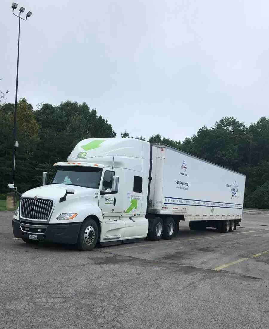 White semi-truck with a long trailer parked in an empty lot, trees in the background in a parking area.