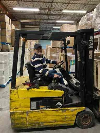 Man in a striped sweater and cap sits in a yellow forklift in a warehouse aisle, surrounded by stacked boxes.